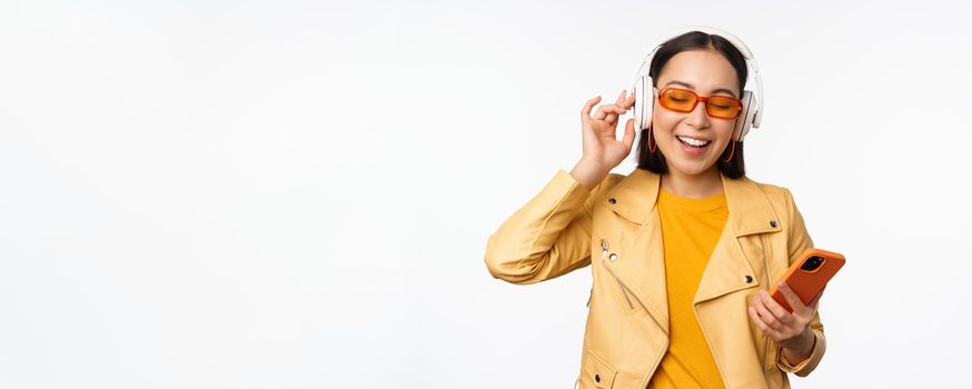 Happy stylish asian woman in sunglasses, listening music in headphones, holding smartphone, singing and dancing, standing over white background.