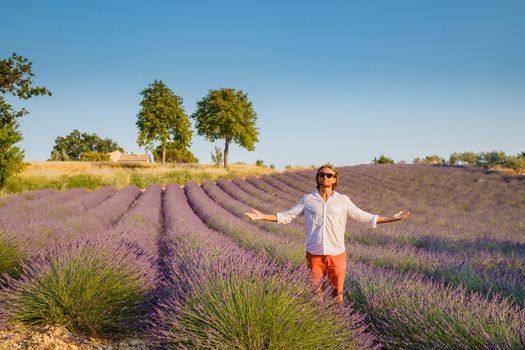 The handsome brutal man with long brunette hair poses in the field of lavender in provence near Valensole, France, clear sunny weather, in a rows of lavender, red shorts, white shirts, blue sky. High quality photo