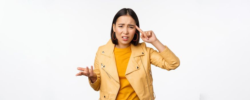 Annoyed asian woman roll finger near temple and staring frustrated, scolding someone stupid or crazy, standing over white background.