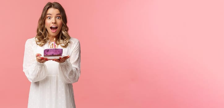 Holidays, spring and party concept. Happy cheerful good-looking blond woman celebrating birthday, holding piece cake with lit candle, making wish, look amused standing pink background.