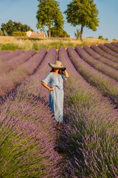 The beautiful young girl in a blue dress walks across the field of a lavender, long curly hair, smile, pleasure, mountains on background, a house of the gardener, trees, perspective of a lavender. High quality photo