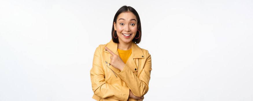 Portrait of beautiful asian woman express intereest and excitement about advertisement, store discount, pointing finger left, looking amazed, standing over white background.