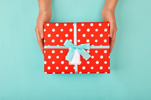 Hands give a red polka dot gift box with a card and a bow on isolated pastel blue-turquoise background top view