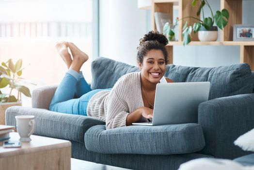 Full length portrait of a happy young woman using her laptop while laying on the sofa at home.