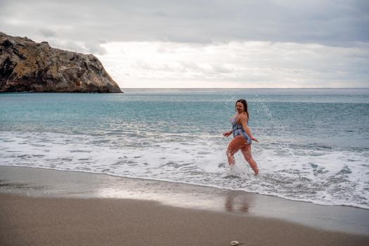 A plump woman in a bathing suit enters the water during the surf. Alone on the beach, Gray sky in the clouds, swimming in winter