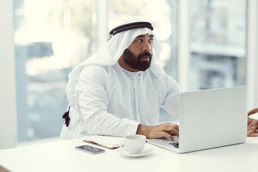 Cropped shot of a young businessman dressed in Islamic traditional clothing working on his laptop while sitting in the office.