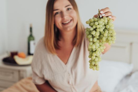Young Caucasian Woman Holding Bunch of Grapes in Hand While Sitting in Hotel Room, Tray of Fruit and Bottle of Wine Stands on Background
