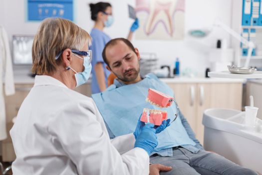 Senior stomatologist woman with face mask holding jaw plastic model explaining oral hygiene to man patient during dentistry examination in stomatological office room. Stomatology concept