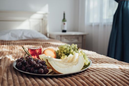 Tray of Fruit Lies on the Bed in Hotel Room, Bottle of Wine Stands on the Background