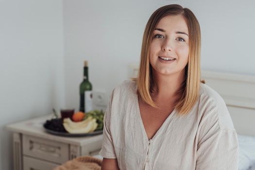 Portrait of Young Caucasian Woman Sitting in Hotel Room, Tray of Fruit and Bottle of Wine Stands on Background