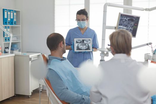 Specialist orthodontist nurse wearing face mask showing teeth radiography to patient discussing medical surgery during stomatology examination in dental office. Sick man having oral infection