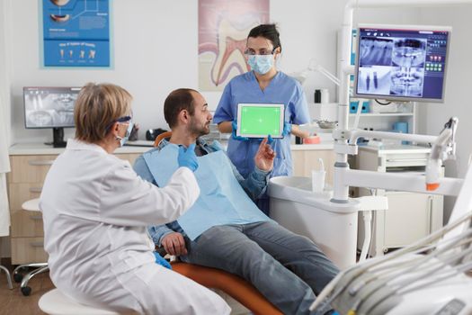 Assistant woman holding mock up green screen chroma key tablet with isolated display discussing oral hygiene with man patient during stomatological consultation in dental office. stomatology concept
