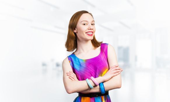 Happy woman standing with folded arms. Elegant good looking lady wears bright dress and bracelets. Joyful redhead girl with perfect teeth smile on blurred office background. Flirty student posing