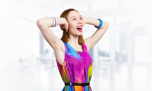 Redhead beautiful girl closing her ears with hands. Charming lady in bright colorful dress and bracelets on blurred office background. Portrait of happy young woman looking up aside with open mouth