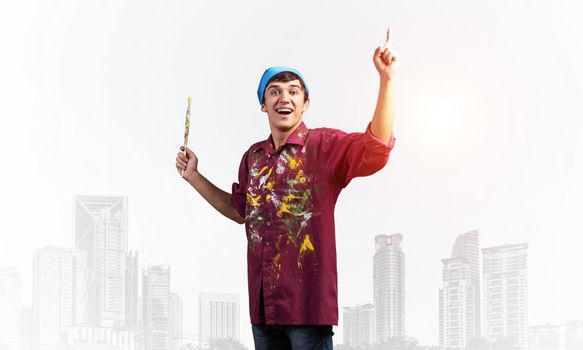 Young emotional artist gesturing with paintbrush. Happy painter in shirt and bandana standing on background modern office buildings. Creative hobby and artistic occupation concept with copy space