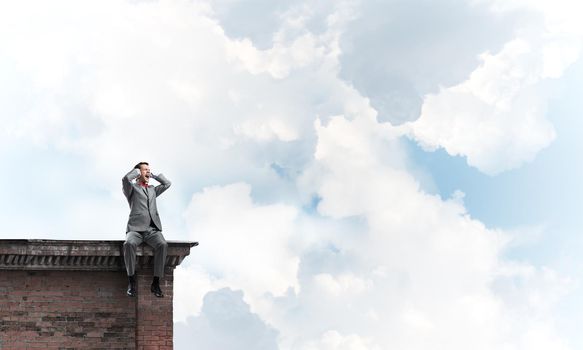 Frustrated businessman sitting on building top and closing ears with hands