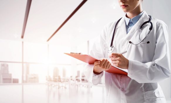 Close up of confident female doctor in white medical uniform making notes into notebook while standing inside of bright hospital building. Medical industry concept