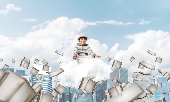 Young little boy keeping eyes closed and looking concentrated while meditating on cloud among flying books with cityscape view on background.