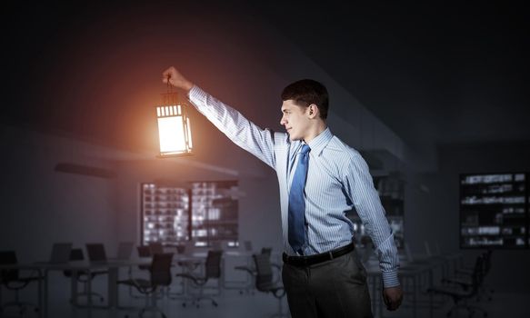 Confident businessman holding glowing lantern on background office interior. Young man in shirt and tie looking for something in dark room. Smiling business person lighting his way with lantern.