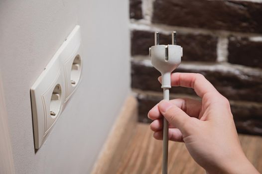 A female hand inserts white electric plug into a euro-format socket close-up.