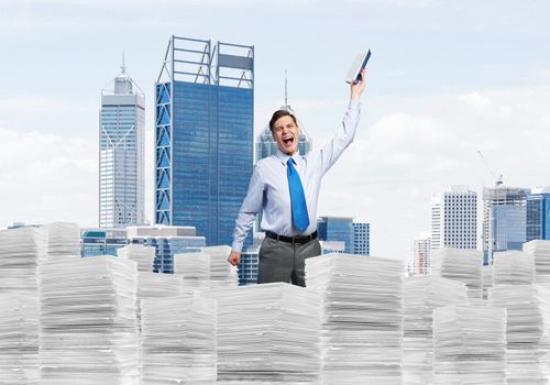 Attractive businessman keeping hand with book up while standing on pile of paper documents with cityscape on background. Mixed media.