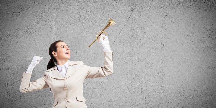 Beautiful woman holding trumpet brass overhead. Young smiling businesslady in white business suit and gloves posing with music instrument on grey wall background. Business concept with musician