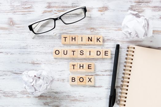 Think outside the box quote with letters on wooden cubes. Still life of office workspace. Flat lay wooden desk with crumpled paper, glasses and spiral notebook. Business motivation and inspiration.