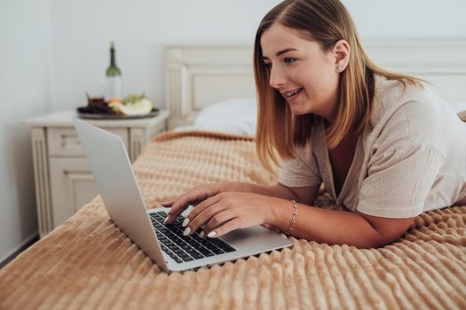 Young Caucasian Woman Laying on Bed in Hotel Room and Working on Laptop, Freelancer at Work