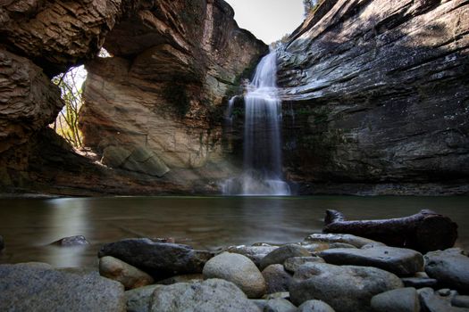 Rays of sunlight entering through a hole in a rock and a waterfall falling on a lake with stones and a big branch in the foreground in a place called Foradada