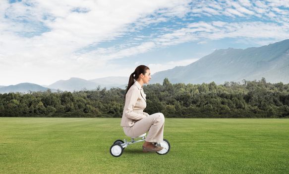 Beautiful young woman riding children's bicycle on green grass. Businesslady in white business suit cycling small bike outdoor. Professional career start concept. Beginner level metaphor.