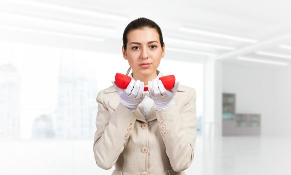 Smiling young woman holding retro red phone. Call center operator in white business suit posing with telephone in light office interior. Hotline telemarketing. Business assistance and consultation.