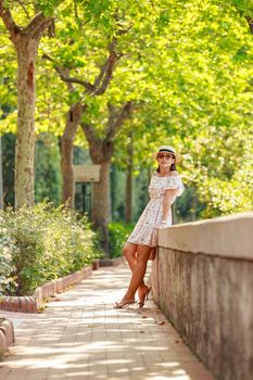 The bright beautiful girl in a light dress and hat walks along alley of trees in Monaco in sunny weather in the summer, handbag in hand. High quality photo