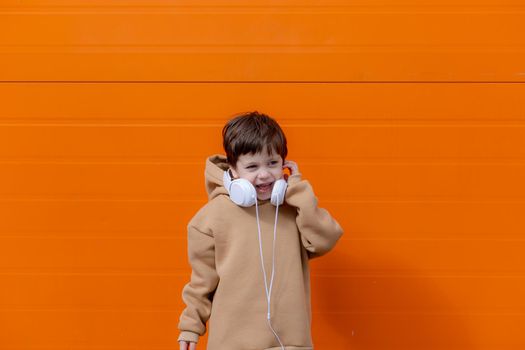 A boy listens to music with headphones on the background of an orange wall . Copy space. Mockup. An article about modern children. An article about modern music. Article about the choice of headphones. Urban lifestyle