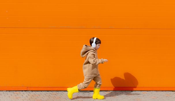 A boy listens to music with headphones on the background of an orange wall . Copy space. Mockup. An article about modern children. An article about modern music. Article about the choice of headphones. Urban lifestyle