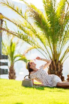 a bright beautiful girl in a light dress and hat lies on the grass under a palm tree in Monaco in sunny weather in summer, streets of old town of Monaco. High quality photo