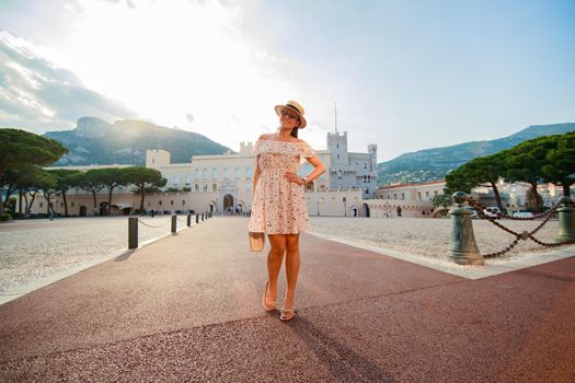 a bright beautiful girl in a light dress and hat walks along the streets of old town of Monaco in sunny weather, a residential area of principality and walking people in the background. High quality photo