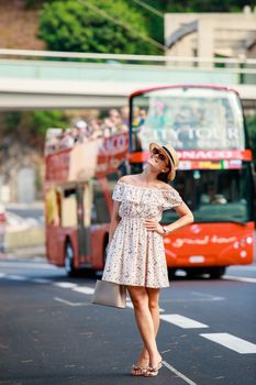 a bright beautiful girl in a light dress and hat walks along the streets of Monaco in sunny weather in the summer, tourist red bus in the background. High quality photo
