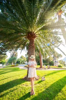 The bright beautiful girl in a light dress and hat standing under a palm tree of Monaco in sunny weather in summer. High quality photo