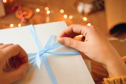 Girl's hands tie blue ribbon bow on white gift box. Garland lights and decorations in background. Surprise for Birthday or Christmas. New Year's mood.