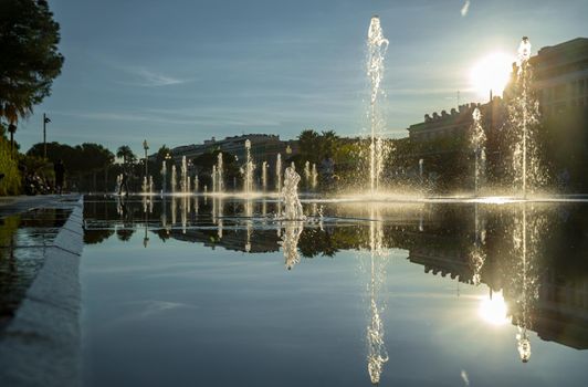 France, Nice, Famous place of Nice, square of fountain, mirror of water, playing kids, walking couples, a couple of old people are basking in the sunset, sun reflection, passing tram. High quality photo