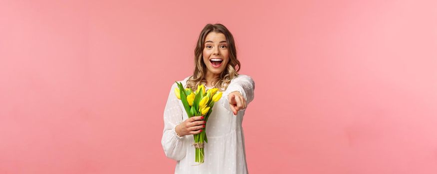 Holidays, beauty and spring concept. Portrait of excited happy and upbeat young blond girl in white dress, holding bouquet of yellow tulips, pointing finger at camera amazed, pink background.