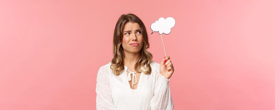 Spring, happiness and celebration concept. Close-up of skeptical and doubtful good-looking blond girl holding commend cloud near head as if having thought, grimacing and squinting from dislike.