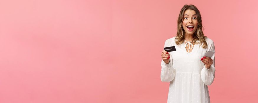 Portrait of excited and amused, happy smiling caucasian woman making order internet, shopping online with mobile phone, enter credit card, billing info, look camera amazed, pink background.