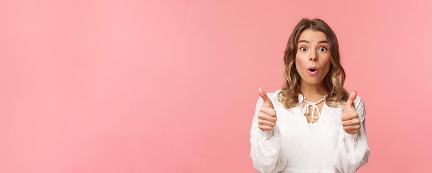 Close-up portrait of impressed blond girl showing thumbs-up look astonished and disbelief as processing fascinating performance she saw, leave positive review, standing pink background.