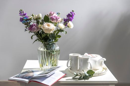 Bouquet of hackelia velutina, purple and white roses, small tea roses, matthiola incana and blue iris in glass vase is on the white coffee table with two tall cups for tea. Grey wall