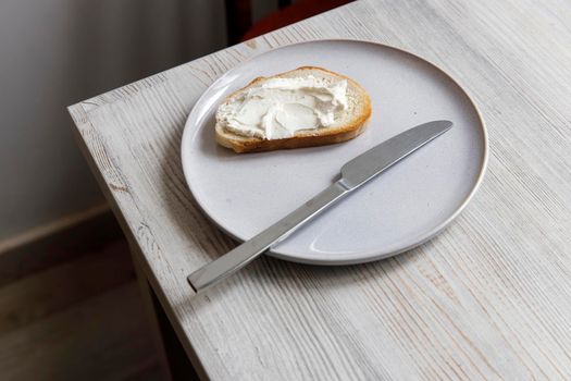 A piece of bread with curd cheese spread on it on a white ceramic plate with a knife on the beige table.