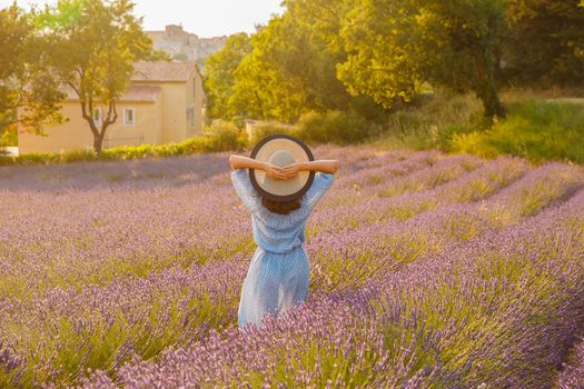 The beautiful young girl in a blue dress and cap walks across the field of a lavender, long curly hair, smile, pleasure, a house of the gardener in the background, trees, perspective of a lavender. High quality photo