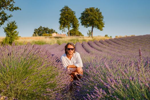 The handsome brutal man with long brunette hair sits on field of lavender in provence near Valensole, France, clear sunny weather, in a rows of lavender, red shorts, white shirts, blue sky. High quality photo