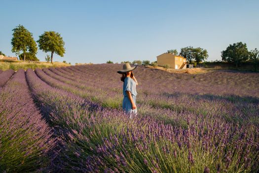 The beautiful young girl in a blue dress and cap walks across the field of a lavender, long curly hair, smile, pleasure, a house of the gardener in the background, trees, perspective of a lavender. High quality photo