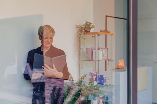 portrait through a window of a smiling mature professional Beautician with blonde hair, dressed in black work uniform, checking email on her Tablet. owner in her small beauty salon looking at laptop. Work from home, enterprising woman, active small business owner. Relaxed atmosphere and soft lighting from window, natural light, tablet. horizontal.